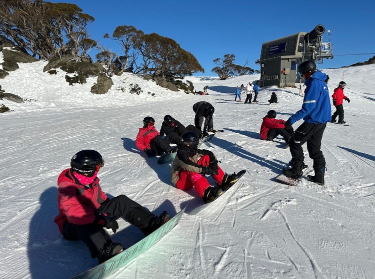 Photo of a sunny day in the snow with students in red jackets sitting down and an instructor in a blue jacket standing near them